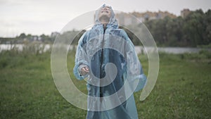 Camera approaches to joyful Caucasian boy in raincoat having fun on rainy summer day outdoors. Portrait of cheerful cute