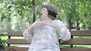 Camera approaches to senior woman sitting on bench holding chest. Portrait of Caucasian retiree having symptoms of heart