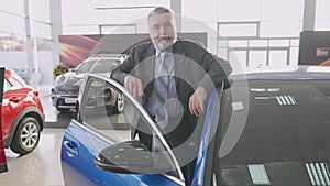 Camera approaches to smiling happy senior man standing at open blue car in showroom. Portrait of confident cheerful