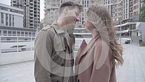 Camera approaches to young man kissing woman on windy urban street. Side view portrait of young Caucasian couple having