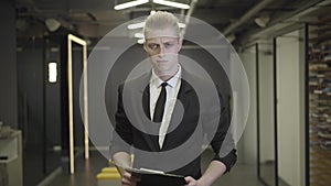 Camera approaching serious redhead man in elegant black formal suit standing in open space corridor. Male office worker