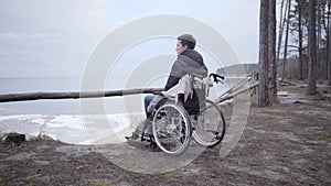 Camera approaching to lonely sad Caucasian boy in wheelchair looking away. Young crippled man spending autumn day alone