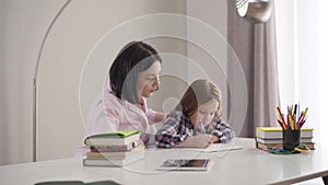 Camera approaching to tired caucasian mother yawning as helping daughter to do homework. Cute brunette schoolgirl