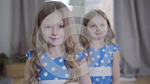 Camera approaching to two beautiful little Caucasian girls posing indoors. Brunette twin sisters in similar dresses