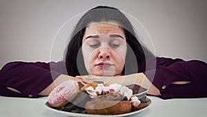 Camera approaching to upset fat Caucasian woman sitting in front of sweets and licking lips. Obese girl looking at tasty