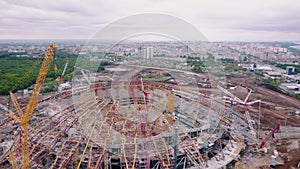 Camera is flying over large sportive arena under construction with city panorama in background