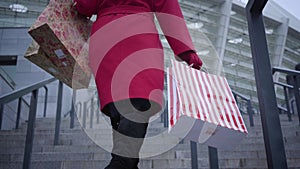Camera following unrecognizable stylish lady in red coat and black boots walking up the stairs in city with shopping