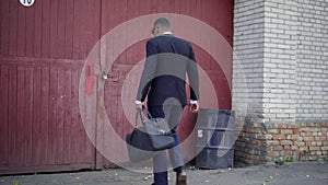 Camera follows African American businessman with bag walking to abandoned industrial slum and knocking in big red wooden