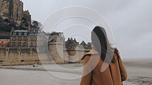 Camera follows beautiful happy tourist woman walking close to epic Mont Saint Michel castle on overcast day slow motion.