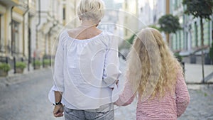 Camera follows carefree Caucasian grandmother and granddaughter strolling on sunny city street and chatting. Back view