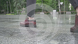 Camera follows footsteps of elegant young Caucasian man. Close-up of male feet in boots walking on pavement along urban