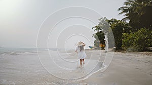 Camera follows happy beautiful little 5-7 year old girl in big straw hat running along atmospheric tropical ocean beach.