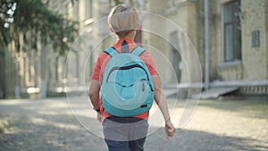 Camera follows little blond schoolboy waving to friends and marching to school in sunlight. Happy Caucasian boy walking