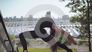 Camera follows movement of young slim Caucasian woman bending backwards at sunrise with urban city at the background
