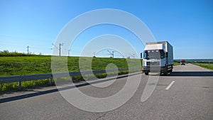 Camera follows to truck with cargo trailer driving on highway transporting goods at summer day. White lorry riding