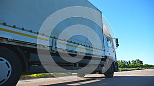 Camera follows to truck with cargo trailer driving on highway and transporting goods at sunny day. White lorry speeding