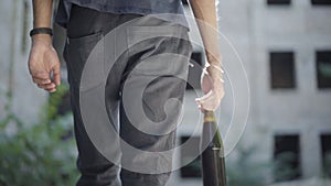 Camera follows unrecognizable young man walking on urban with beer bottle. Back view of alcoholic strolling in sunlight