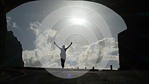 Camera follows a woman silhouette walking out from tunnel towards the sunlight. Burst of bright light. Raising arms in