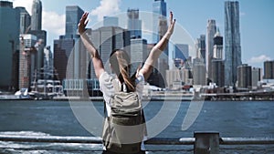 Camera follows young happy traveler girl with backpack, jumping with arms wide open at Manhattan skyline in New York.