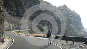 Camera follows young woman with backpack walking from a tunnel towards a cliff edge raising arms in air.