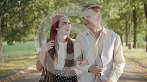 Camera moves up along happy young couple walking and chatting outdoors in sunlight. Portrait of confident relaxed