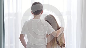 Camera moving away from Caucasian girl and boy standing together in front of the window. Girl`s head is on boy`s