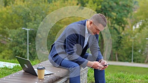 Camera moving forward to handsome man in suit sitting outside in nature browsing online on smartphone. Happy young male