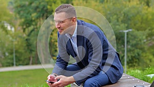Camera moving forward to handsome man in suit sitting outside in nature browsing online on smartphone. Happy young male