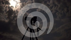 Camera on tripod under night cloudy sky with moon