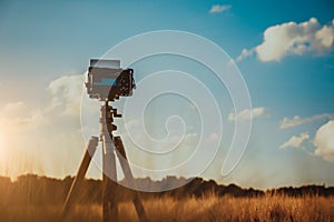 Camera on tripod in wheat field, silhouette on sunset sky with whie moving clouds in background