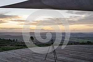 Camera tripod on wooden platform in natural landscape at sunset.