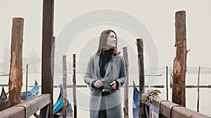 Camera zooms out on happy smiling tourist woman with camera taking photos on romantic Venice gondola pier with red roses