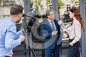 cameraman and news reporter with microphone interviewing businessman near office