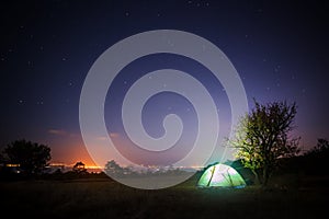 Camp tent stands on glade under starry night sky and shines from