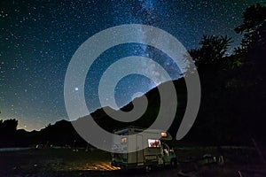 Camper van under panoramic night sky in the Alps. The Milky Way galaxy arc and stars over illuminated motorhome. Camping freedom