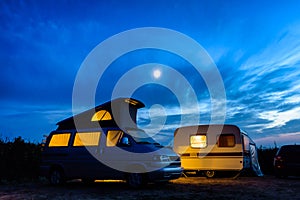 A campervan and a caravan illuminated from inside at nightfall with the moon glowing in a stormy sky.