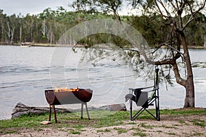 Campfire in a half barrel fire pit and empty campchair on the lake