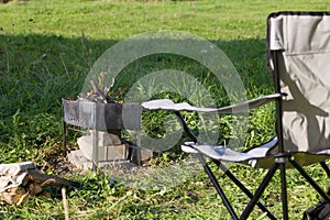 Camping chair and metal grill with a light fire. They are standing in a green meadow. Autumn picnic
