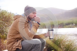 Camping, cold and man with coffee by lake in nature for adventure, travel and outdoor vacation. Holiday, morning and