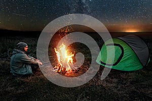 Camping man sits by the fire at night against the background of the starry sky. The concept of travel, tourism, camping