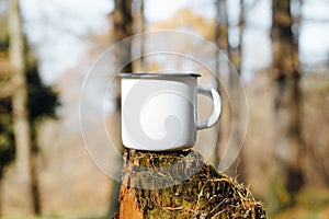 Camping metal white mug mock up standing on tree stump in woods outdoors. Enamelled balnk cup with empty space