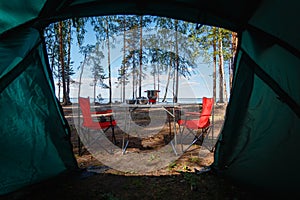 Camping in the middle of the forest, taken from inside the tent. View of the folding table and chairs with gas stove in the backgr