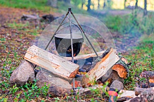 Camping outdoors. Cooking bowler hat hung on tripod over burning fire on the background of grass