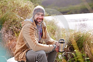 Camping, portrait and man with coffee by lake in nature for adventure, travel and outdoor vacation. Holiday, smile and