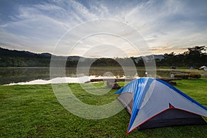 Camping tent on green grass beside lake with foggy over forest during sunrise