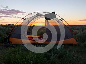 Camping tent with solar charger in quiet grassy meadow while the sunset peers through mesh screen. A peaceful campsite for the eve