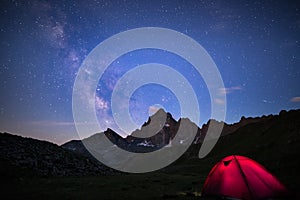 Camping under starry sky and milky way at high altitude on the Alps. Illuminated tent in the foreground and majestic mountain peak