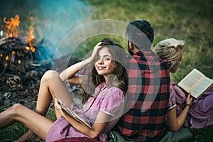 Camping in wilderness. Turn back guy looking at fire while two beautiful girls read book. Smiling brunette with braces