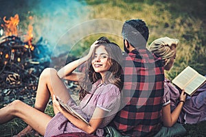 Camping in wilderness. Turn back guy looking at fire while two beautiful girls read book. Smiling brunette with braces