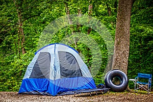 Camping in the woods - Basic blue tent with simple bed outside and tube floaty proped against tree by camp chair - lush foliage